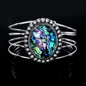 May include: A silver cuff bracelet featuring a large, oval-shaped abalone shell inlay with iridescent blue, green, and purple patterns. The centre stone is framed by a decorative scalloped silver border, and the band consists of four thin wires.