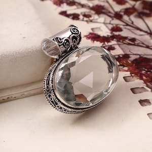 May include: An oval-shaped pendant featuring a large, clear, faceted gemstone set in a decorative silver frame. The metal setting displays intricate, dark-oxidized scrollwork patterns along the edges and a matching ornate bail at the top.