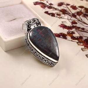 May include: A silver-coloured pendant with a dark blue and red gemstone in a teardrop shape. The metal frame has an intricate scrollwork design. The pendant is presented in a white jewellery box. The gemstone is approximately 3 cm long.