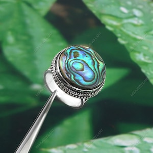 May include: A silver ring featuring a large, oval abalone shell cabochon with iridescent blue, green, and purple hues. The ring has a detailed, beaded border and a simple, polished band. The ring is held by a pair of silver tweezers.