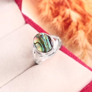 May include: A silver ring featuring an oval-shaped abalone shell inlay. The iridescent shell displays swirling patterns of green, blue, and purple hues. The metal band is smooth and polished, securely holding the cabochon stone in a bezel setting.