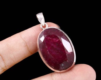 Può includere: Un pendente di rubino con un'ansa color argento. Il grande rubino di forma ovale è di colore rosso intenso ed è sfaccettato per catturare la luce. Il pendente è tenuto tra due dita su sfondo nero.