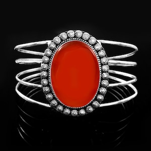 May include: A silver cuff bracelet featuring a large, smooth, oval-shaped orange stone at the center. The stone is surrounded by a decorative border of small, textured silver beads. The band consists of four thin, parallel silver wires.