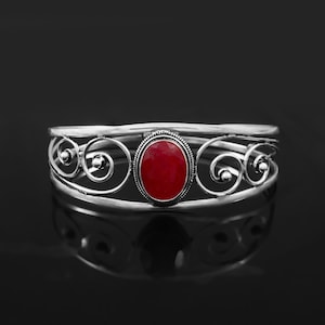 May include: A silver cuff bracelet featuring a central oval-shaped red gemstone. The metal band is designed with openwork scroll patterns on both sides of the stone, creating a symmetrical appearance with polished silver wire detailing.