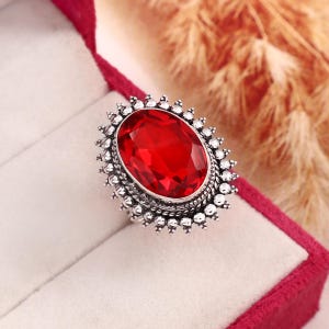 May include: A silver ring featuring a large, oval-cut, vibrant red gemstone at its centre. The setting is crafted with intricate metalwork, including a beaded border and a twisted rope design surrounding the stone, creating a detailed frame.