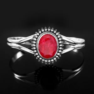 May include: A silver cuff bracelet featuring a central oval-shaped red gemstone. The stone is surrounded by a decorative beaded metal border. The band splits into two smooth, polished silver arms on each side of the central setting.