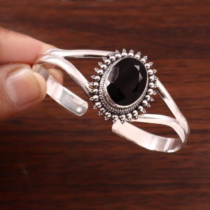 May include: A silver cuff bracelet featuring a central oval-shaped black gemstone. The stone is surrounded by a decorative beaded metal border. The bracelet band consists of two split silver wires that meet at the sides to form a single band.