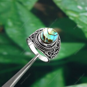 May include: A silver ring with an oval, iridescent abalone shell center stone. The ring has an ornate, textured band with a braided design and decorative filigree elements. The abalone shell displays shades of blue, green, and brown.