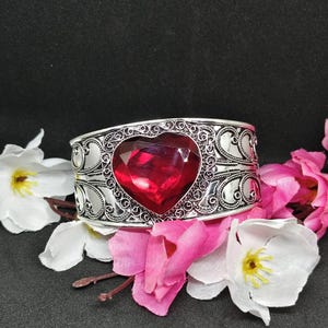 May include: A silver-toned cuff bracelet featuring an intricate filigree pattern and a large, faceted, heart-shaped red gemstone centerpiece. The metalwork displays detailed scrollwork surrounding the central stone, creating a decorative band.