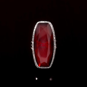 Op de afbeelding: Een ring met een grote, langwerpige, gefacetteerde dieprode edelsteen gezet in een gestructureerd zilverkleurig metalen frame. De steen heeft een geometrisch, veelzijdig oppervlak dat licht reflecteert, met een kralenrand rondom de zetting.