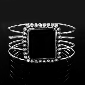 Könnte beinhalten: Ein silberfarbenes Armband mit einem großen, rechteckigen, glatten schwarzen Stein in der Mitte. Der Stein ist von einem dekorativen Rand aus kleinen, strahlenförmigen Metalldetails umgeben. Das Armband besteht aus vier dünnen, parallelen Silberdrähten.