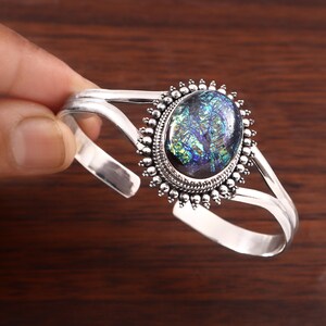 May include: A silver cuff bracelet featuring a central oval-shaped stone with iridescent blue, green, and purple flecks. The stone is surrounded by a decorative beaded metal frame, and the band consists of two split silver arms that meet at the sides.