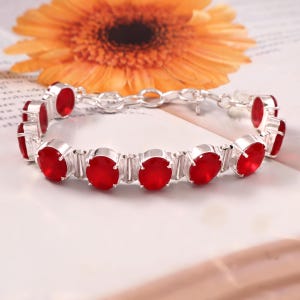 May include: A silver-toned link bracelet featuring a series of round, vibrant red stones set in prong settings. The bracelet includes a circular clasp mechanism and is constructed with alternating metal links between each of the red circular stones.