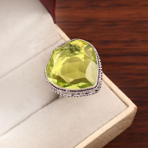 Puede incluir: Un anillo con una gran piedra preciosa de color verde lima, facetada y en forma de corazón, engastada en una banda de plata oxidada decorativa con intrincados detalles de metal con cuentas y filigrana en los bordes del engaste.
