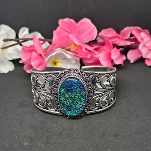 Puede incluir: Una pulsera tipo brazalete de tono plateado con una pieza central ovalada de superficie texturizada iridiscente en tonos verdes y azules. La banda metálica está decorada con intrincados patrones de filigrana que enmarcan la piedra central.