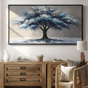 Puede incluir: Una obra de arte enmarcada que representa un árbol con flores azules y blancas. El tronco y las ramas oscuras del árbol contrastan con el fondo claro. La obra de arte se exhibe sobre una cómoda de madera, con una lámpara y una silla visibles.