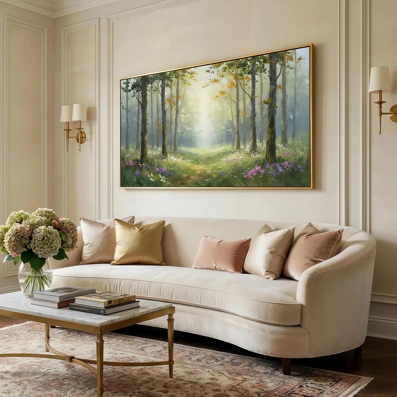 May include: A rectangular landscape painting in a gold frame depicting a sunlit forest path with tall trees, green foliage, and patches of purple and white wildflowers. The artwork is displayed on a wall above a curved cream-colored sofa.
