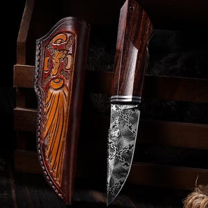 Puede incluir: Un cuchillo de hoja fija con mango de madera oscura y hoja de acero damasco con patrón, acompañado de una funda de cuero marrón con detalles en relieve y bordes cosidos, presentado en una orientación vertical.