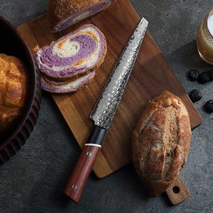 May include: A bread knife with a serrated blade and a wooden handle lies on a wooden chopping board. Sliced purple and white bread is next to the knife. A loaf of bread and a bowl of bread are also in the image.