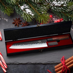 Op de afbeelding: Een Japanse koksmes met een donker, getextureerd lemmet en een houten handvat, gepresenteerd in een zwarte doos met een rode binnenkant. Een kleine slijpsteen is inbegrepen. Feestelijke vakantiedecoraties.