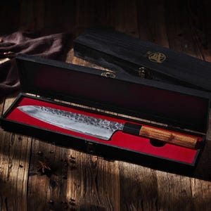 May include: A Japanese-style chef's knife with a wooden handle and a textured blade, presented in a black wooden box with a red velvet interior. The box is open, revealing the knife's full length.