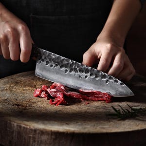 May include: A large, textured metal chef's knife cutting raw red meat on a wooden cutting board. The knife has a dark handle and a blade with a dimpled surface. The meat is freshly cut and appears to be beef.