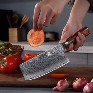 May include: A stainless steel cleaver with a wooden handle is held by a person, slicing a tomato. The blade has a decorative pattern and the word "SAKARI" is visible. Red tomatoes and onions are on a wooden cutting board.
