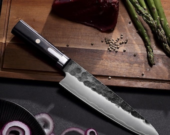 May include: A chef's knife with a dark wooden handle featuring a circular inlay and a silver bolster. The blade has a hammered, textured finish on the upper portion and a smooth, polished edge with a visible wavy temper line along the cutting edge.
