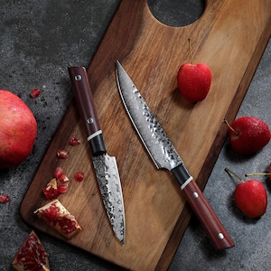 Puede incluir: Dos cuchillos de cocina con mangos de madera oscura y hojas estampadas descansan sobre una tabla de cortar de madera. Manzanas rojas y semillas de granada están esparcidas. Los cuchillos tienen una hoja de metal oscuro con un patrón.