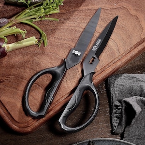 May include: Two black kitchen shears with curved handles and sharp blades, placed on a wooden chopping board. The shears have a distinctive design, with the brand name "Sakari" visible on one blade. Fresh vegetables are in the background.