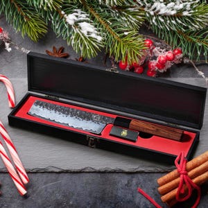 May include: A Japanese-style chef's knife with a hammered blade, presented in a black wooden box with a red interior. The knife has a wooden handle and is accompanied by a small black case. The scene is decorated with Christmas elements.
