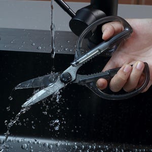 以下が含まれることがあります： 透明なプラスチック製のハンドルが付いたステンレス鋼のキッチンバサミが、流水の下にあります。ハサミにはギザギザのエッジと、ブランド名「SAKARI」が見えるピボットポイントがあります。水滴が暗い背景に対してハサミの周りに散らばっています。