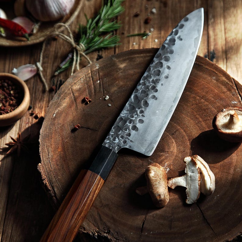 May include: A handcrafted chef's knife with a wooden handle and a textured, silver-colored blade. The knife rests on a wooden cutting board, alongside mushrooms and spices, suggesting a culinary theme.