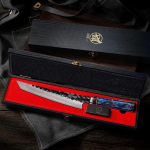 Puede incluir: Un cuchillo de chef de estilo japonés con una hoja estampada y un mango azul y morado, presentado en una caja de madera negra abierta con interior rojo. También incluye un pequeño soporte a juego.