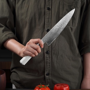 Peut inclure: Un couteau de chef en acier inoxydable avec une lame à motifs et un manche blanc. Le couteau est tenu devant une personne portant une chemise vert foncé. Une tomate est visible au premier plan.