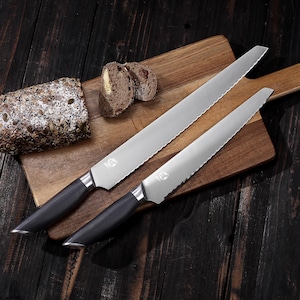 May include: Two serrated bread knives with black handles rest on a wooden cutting board next to a loaf of bread. The knives have silver blades and are designed for slicing bread. The bread is sliced and has seeds on the crust.