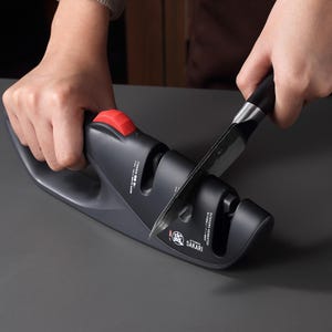 May include: A dark gray knife sharpener with a red button is being used to sharpen a black knife. The sharpener has the brand name "Sakari" printed on it. The knife is being pulled through the sharpener.