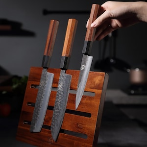 May include: Three Japanese-style kitchen knives with dark wooden handles and textured blades are displayed in a wooden knife block. The knives have a dark metal bolster and a hammered finish on the blades. The knife block is a rich, dark brown color.