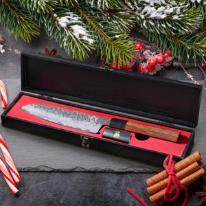 Peut inclure: Un couteau de chef japonais avec un manche en bois et une lame texturée, présenté dans un coffret noir avec un intérieur rouge. Un petit accessoire noir est également présent. La scène est décorée d'éléments festifs.