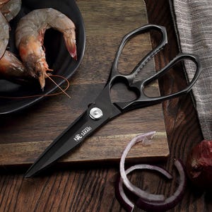 May include: Black kitchen shears with the brand name "Sakari" are displayed on a wooden cutting board. The shears are next to a plate of shrimp and onion slices. The shears have a heart-shaped opening in the handle.