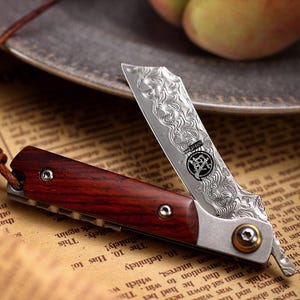 Könnte beinhalten: Ein Klappmesser mit einer gemusterten Stahlklinge und einem dunklen Holzgriff. Die Klinge ist teilweise geöffnet und zeigt ein kompliziertes Design. Das Messer hat einen kleinen, runden, messingfarbenen Verschluss. Der Hintergrund umfasst bedrucktes Papier und einen verschwommenen Teller.