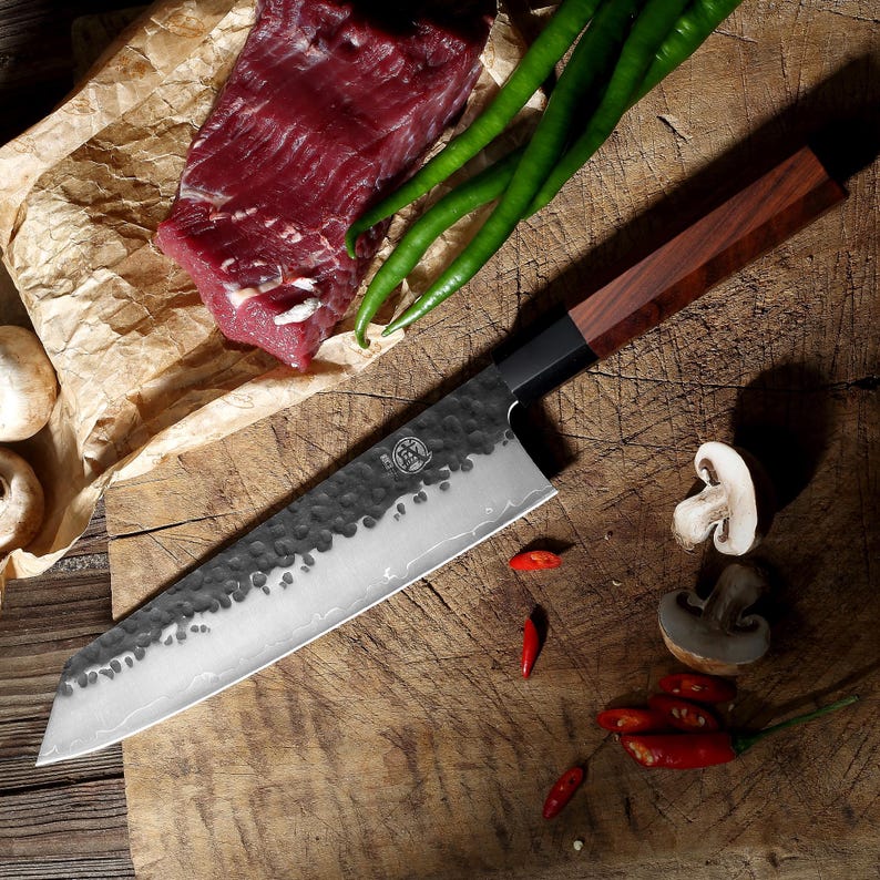May include: A large, stainless steel chef's knife with a dark wooden handle rests on a wooden cutting board. The blade has a textured, hammered finish. Fresh ingredients, including meat, green peppers, mushrooms, and red chilies, are arranged around the knife.