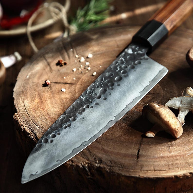 May include: A close-up of a Japanese-style chef's knife with a wooden handle and a textured, gray blade. The knife rests on a wooden cutting board, with mushrooms and spices nearby. The blade is approximately 12 inches long.