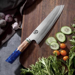Puede incluir: Un cuchillo de chef de acero inoxidable con una hoja estampada y un mango de madera y resina azul. El cuchillo descansa sobre una tabla de cortar de madera, con pepinos en rodajas, tomates y verduras.