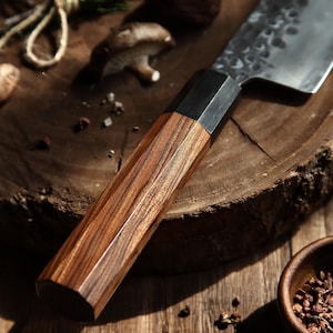 May include: Close-up of a Japanese-style chef's knife with a dark wood handle and a hammered steel blade. The handle has a geometric design. The knife rests on a wooden cutting board, with mushrooms and spices in the background. Culinary tool.