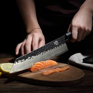 May include: A large, textured stainless steel chef's knife with a black handle is slicing through a piece of salmon on a wooden cutting board. The salmon is bright orange, and a lemon wedge is visible. The knife has a unique, hammered finish.