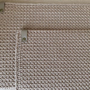 Könnte beinhalten: Zwei beige gehäkelte Tischsets mit Lederriemen und Metallschnappern. Die Tischsets sind rechteckig und haben eine strukturierte Oberfläche. Das kleinere Tischset liegt auf dem größeren. Die Tischsets sind aus weicher Naturfaser gefertigt.