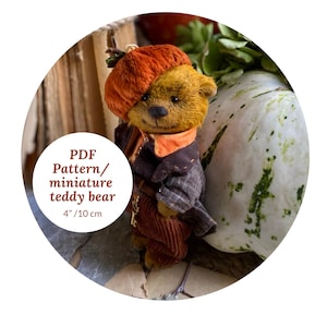 Puede incluir: Un osito de peluche en miniatura, de aproximadamente 10 cm de altura, que lleva un sombrero con forma de calabaza y un atuendo marrón y naranja. El oso tiene una cara detallada y posa frente a una calabaza blanca y otros elementos otoñales. La imagen incluye el texto "PDF Pattern/ miniature teddy bear".