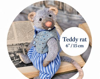 Op de afbeelding: Een grijze pluche rat van 15 cm hoog. De rat draagt een lichtblauw vestje met kleine donkere stippen en drie knopen, gecombineerd met een broek met blauwe en witte verticale strepen. Het heeft een roze neus, zwarte ogen en lange snorharen.