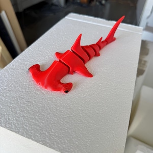 Könnte beinhalten: Ein leuchtend rotes, 3D-gedrucktes Hammerhai-Modell. Der Hai liegt auf einer weißen, strukturierten Oberfläche. Das Modell hat ein detailliertes Design, einschließlich des markanten hammerförmigen Kopfes und der Flossen. Ein auffälliger Dekorationsartikel.